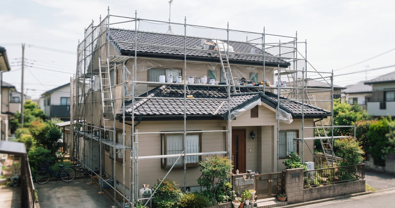戸建て住宅の外観、屋根の周りに足場が組まれ、足場の上に塗料缶や道具が置かれた屋根塗装の準備中の様子